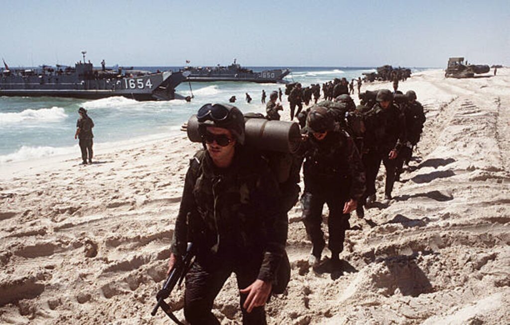 Marines offload from ships onto a beach and walk in lines from landing