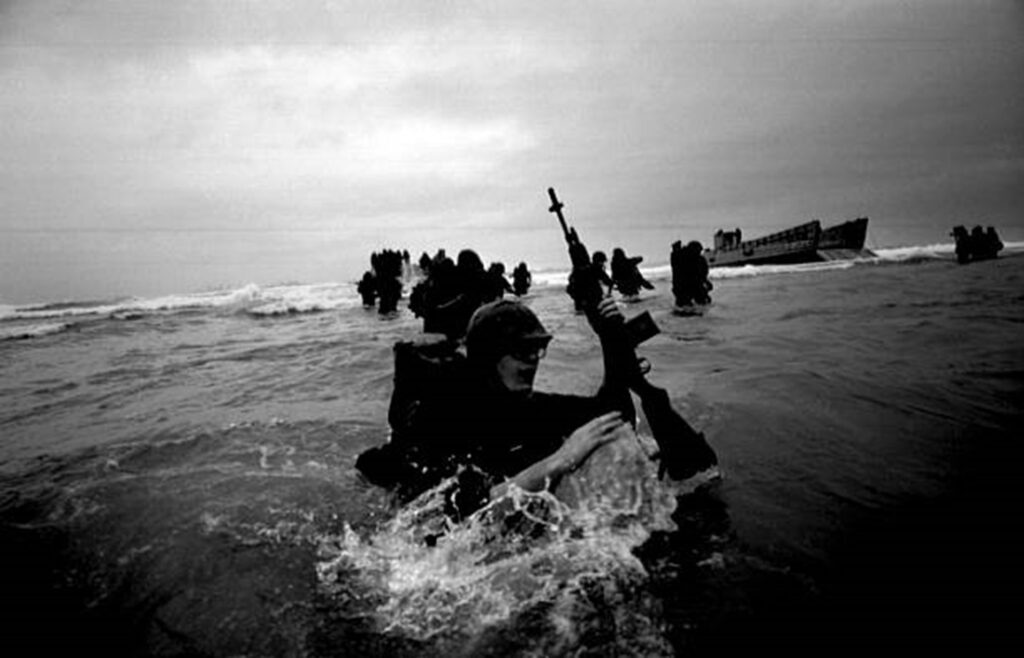 Black and white image of Marines landing on a beach with waves crashing on them.