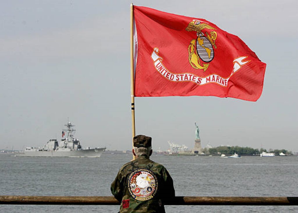 Marine Corps flag with veteran holding it, over looking a war ship as it crosses in front of Elise Island and the Statue of Liberty.